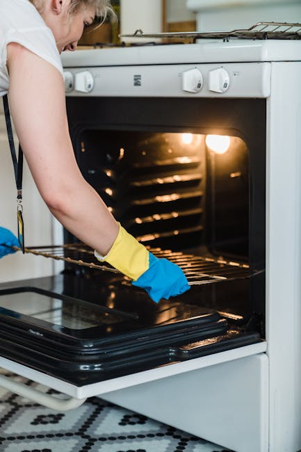 Close-up view of a black, stainless steel oven interior with a person wearing blue gloves and a yellow apron wiping a soapy pink cloth across the oven floor. The surface appears to be non-stick and is