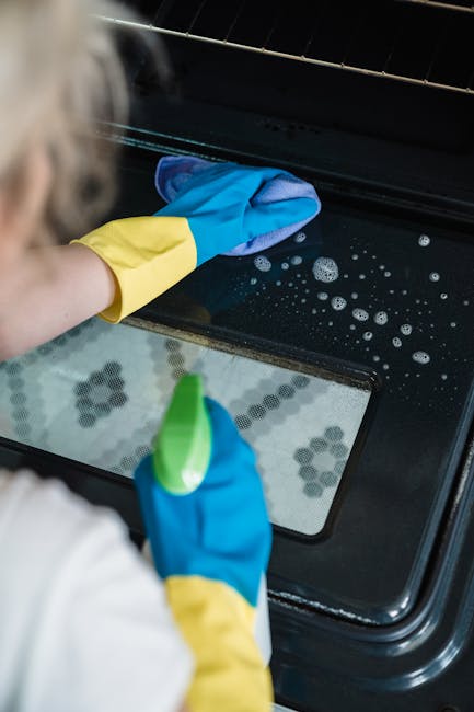 A person wearing yellow and blue cleaning gloves is opening the door of a white oven, situated in a kitchen with patterned black and white tiled flooring. The stove's interior light is on, illuminatin