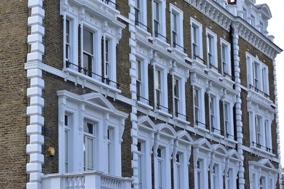 Close-up image of a multi-story red brick residential building facade with numerous white-framed sash windows. The windows are arranged in a grid pattern, with some featuring curtains or blinds visibl