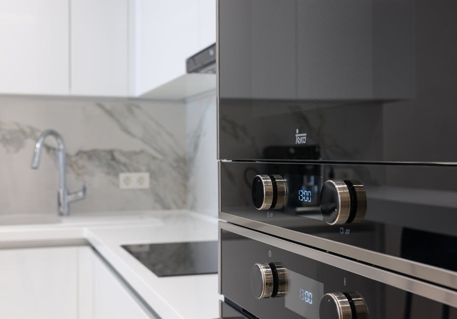 A person wearing blue and yellow rubber cleaning gloves is applying a cleaning solution to the glass surface of an oven door. The oven, with a stainless steel frame, shows soap suds and bubbles on the